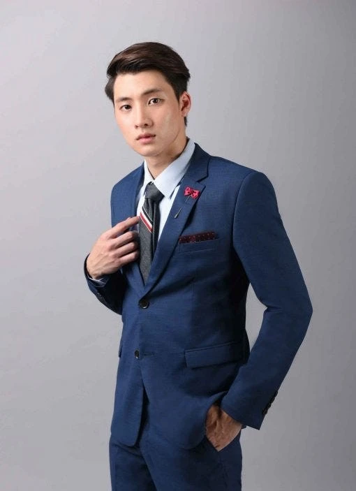 ชายในสูทสีเทาเข้ม สวมเสื้อเชิ้ตขาวและเนคไทสีแดงเข้ม ยืนยิ้ม มือในกระเป๋ากางเกง อยู่หน้าต่างบานใหญ่